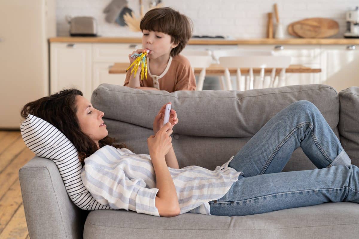 Calm peaceful mom or nanny lying on sofa with mobile phone distracted from noisy loud son drawing attention. Young mother relax at home with smartphone rest after work or parent routine on lockdown