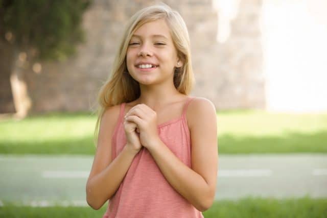 Happy Caucasian little kid girl wearing dress standing outdoors stands against orange studio wall keeps hands on heart, expresses gratitude. Honesty concept.