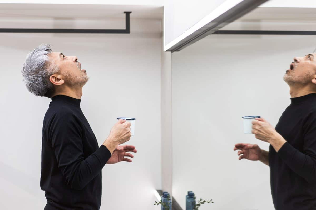 A middle-aged man is gargling in the bathroom with a cup in his hand.