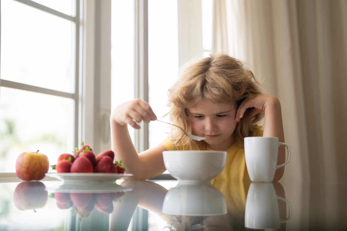 Unhappy little boy child sit at table at home kitchen have no appetite for healthy breakfast. Upset kid refuse to eat cereals with milk. Adorable sad tired kid eating low fat soy milk in bowl cereal.