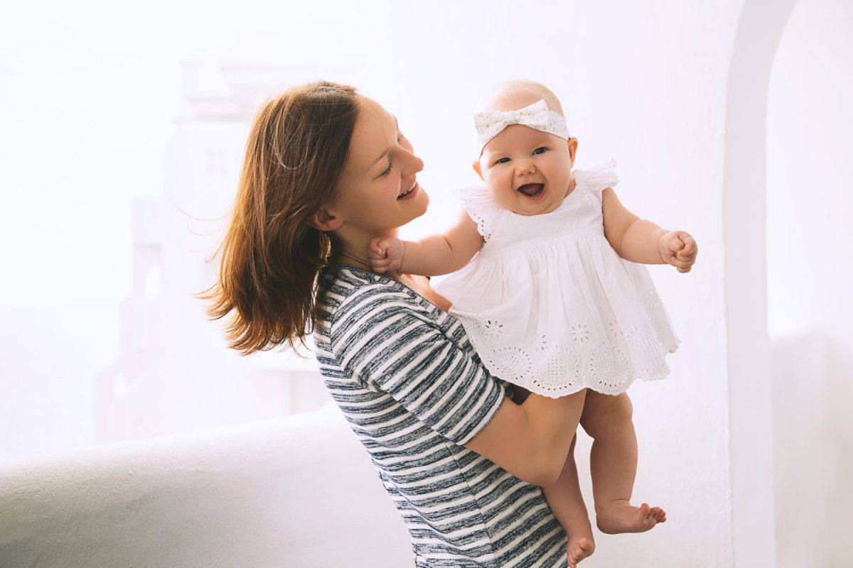 Happy mother and cute 5 months baby girl. Mom with daughter in little white dress, outdoors. Smiling woman and her child on background of white hotel in Greek style on the island of Tenerife, Spain