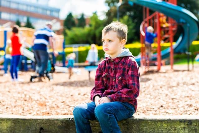 A handsome boy with ADHD, Autism, Asperger Syndrome sitting at the park, scared and apprehensive about playing with the other children, looking nervous and worried