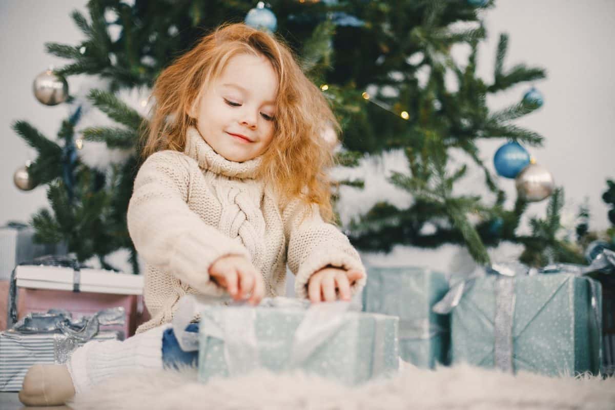 beautiful blonde todler girl opening presents next to the christmas tree