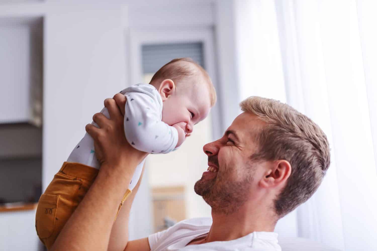 Happy smiling handsome young caucasian dad lifting his loving 6 months old son while standing in living room next to window.