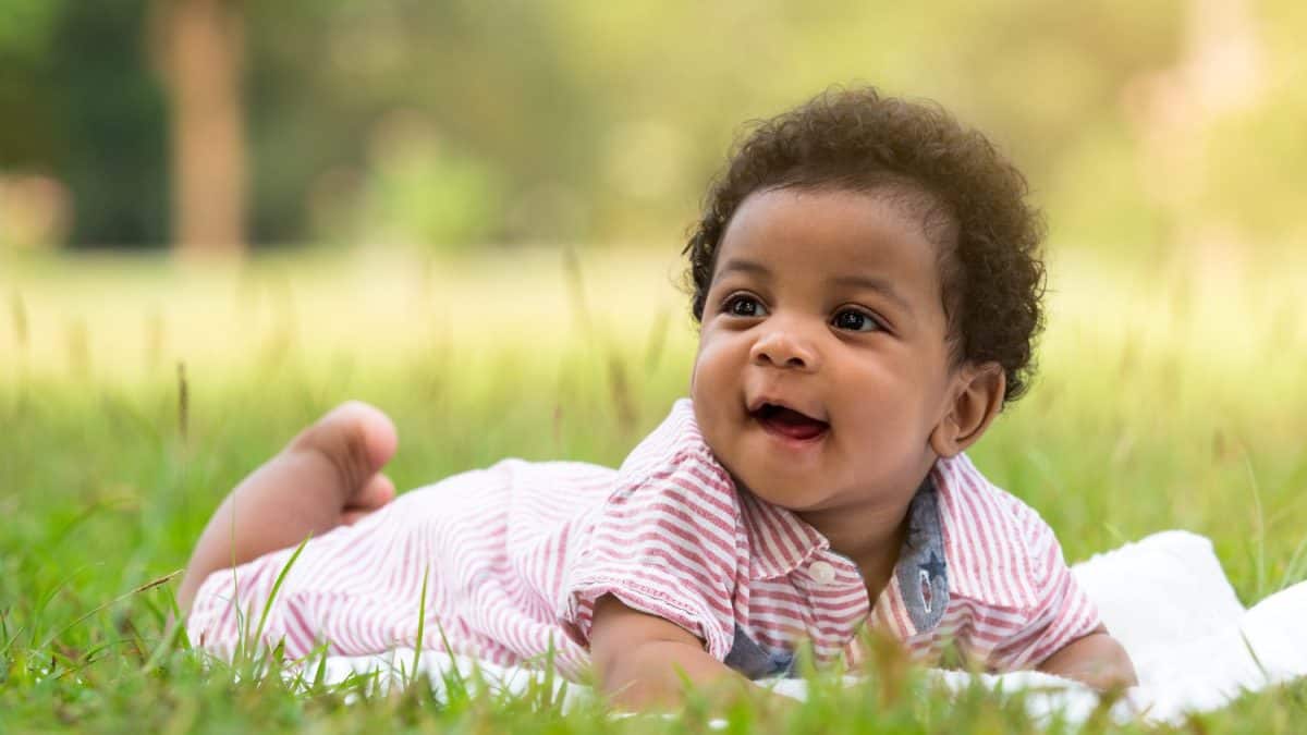 Cute little dark skinned, mix race African baby boy smiling with happiness while lying on the grass in the park.