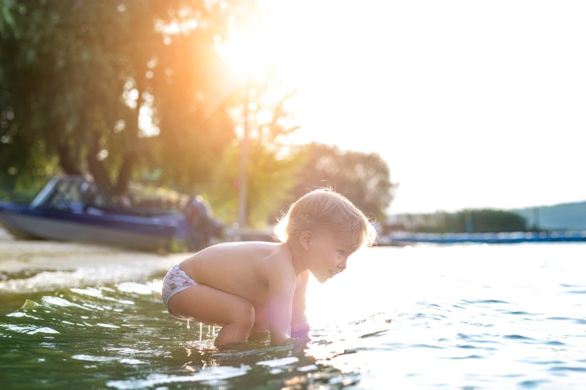 Cute adorable cheerful caucasian little blond toddler boy enjoy having fun playing at lake or river beach water on warm sunset evening time outdoors.. Happy childhood vacation at countryside concept