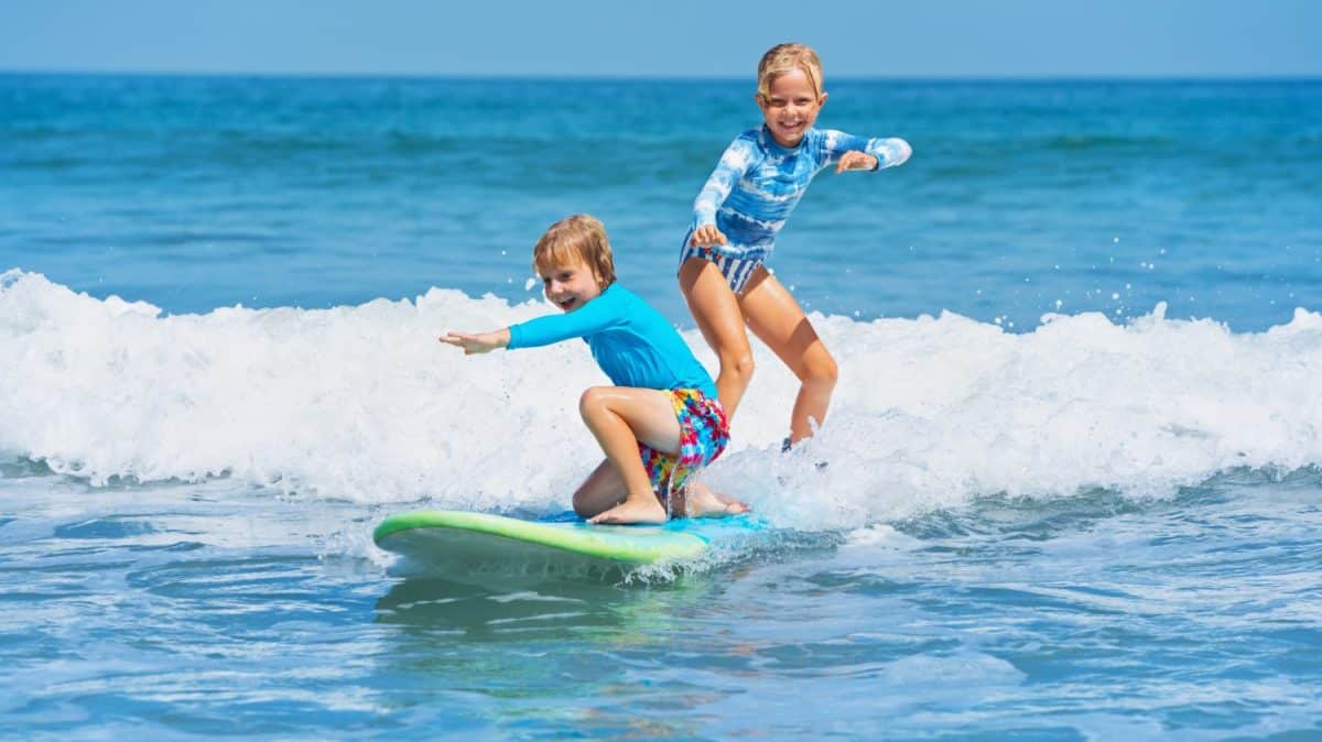 Happy baby boy and girl - young surfers ride with fun on one surfboard. Active family lifestyle, kids outdoor water sport lessons, swimming activity in surf camp. Sea beach summer holiday with child.