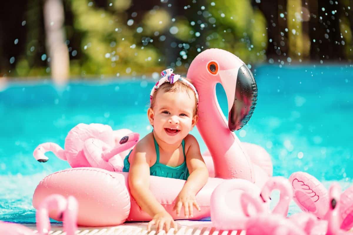 Funny blonde toddler girl swimming in the pool with pink flamingos and swim ring with water splash in the summer