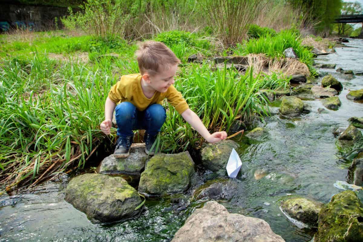 A European boy launches a paper boat in a stream. Children's games by the water. Safety of children in reservoirs.