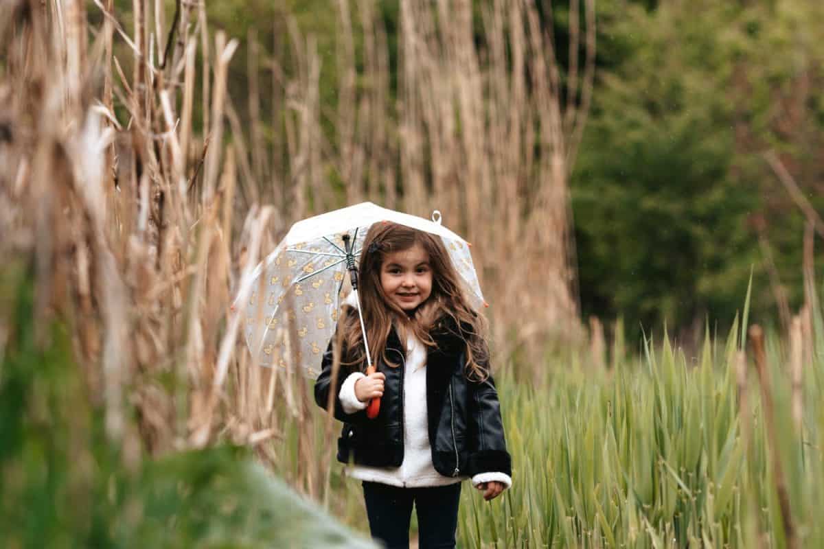 little girl with dark hair and brown eyes dressed in a black jacket rubber boots walks on a rainy day under an umbrella jumps on puddles