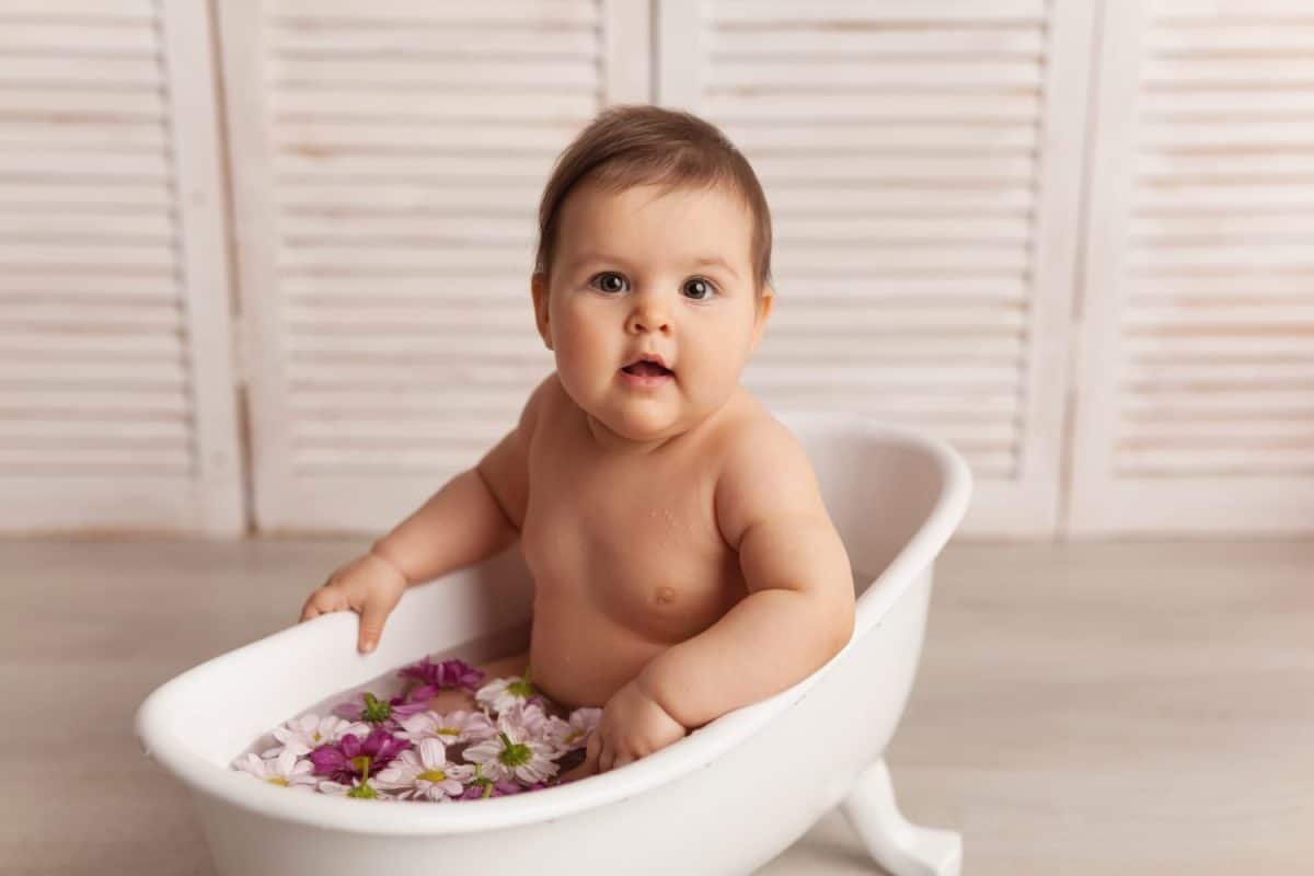 A small European girl with black hair and dark eyes is sitting in a white bathtub in water with flowers and looks surprised