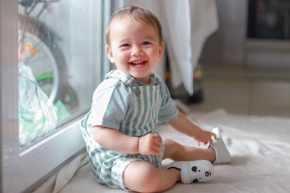 Happy laughing baby boy with teething wearing green dress sitting next to window home.Portrait of mixed race Asian-German infant about one year old joyful with copy space for text.