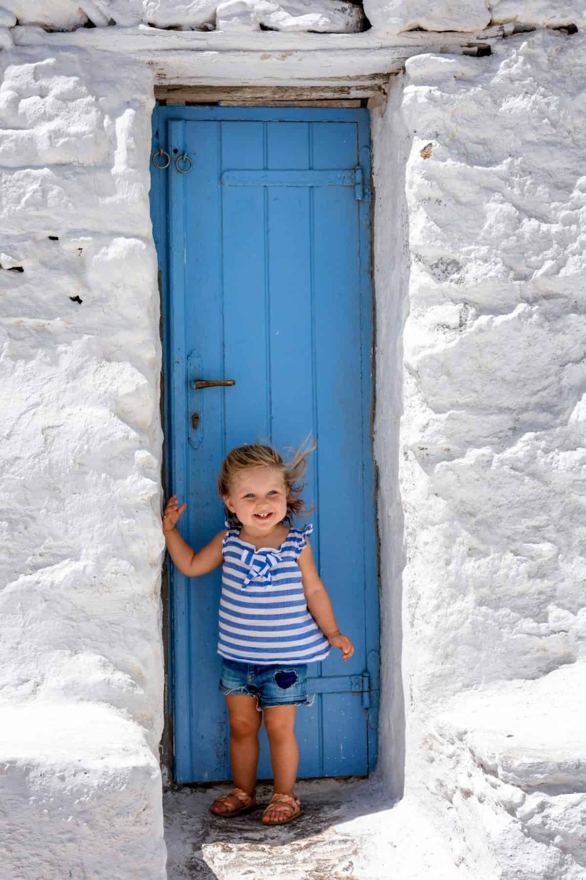 A cute, little baby girl stands on a traditional blue door in front of whitewashed wall on the island of Mykonos, Cyclades, Greece