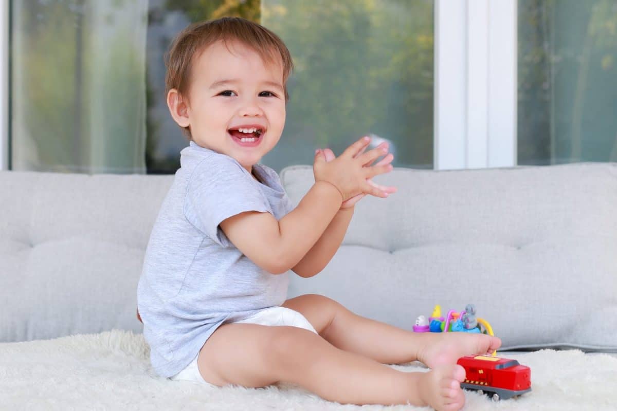 Adorable baby boy about one year old clap his hands while playing with toy at home. Mixed race Asian-German infant in diaper happy and laughing face.