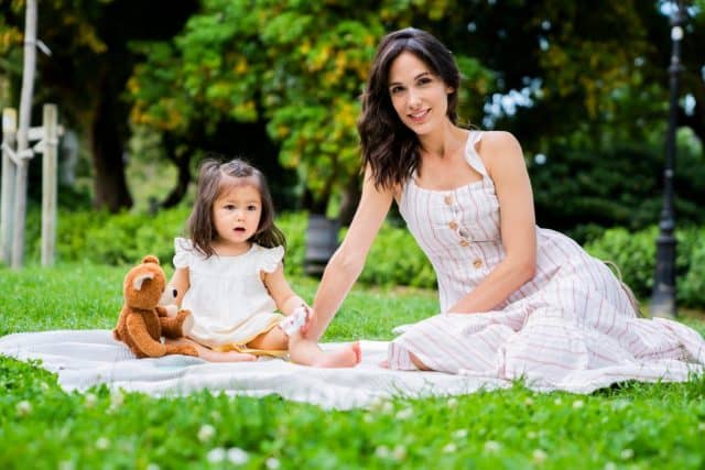 Dark haired cheerful female in striped casual dress having picnic in park with baby girl in white dress with teddy bear and looking at camera