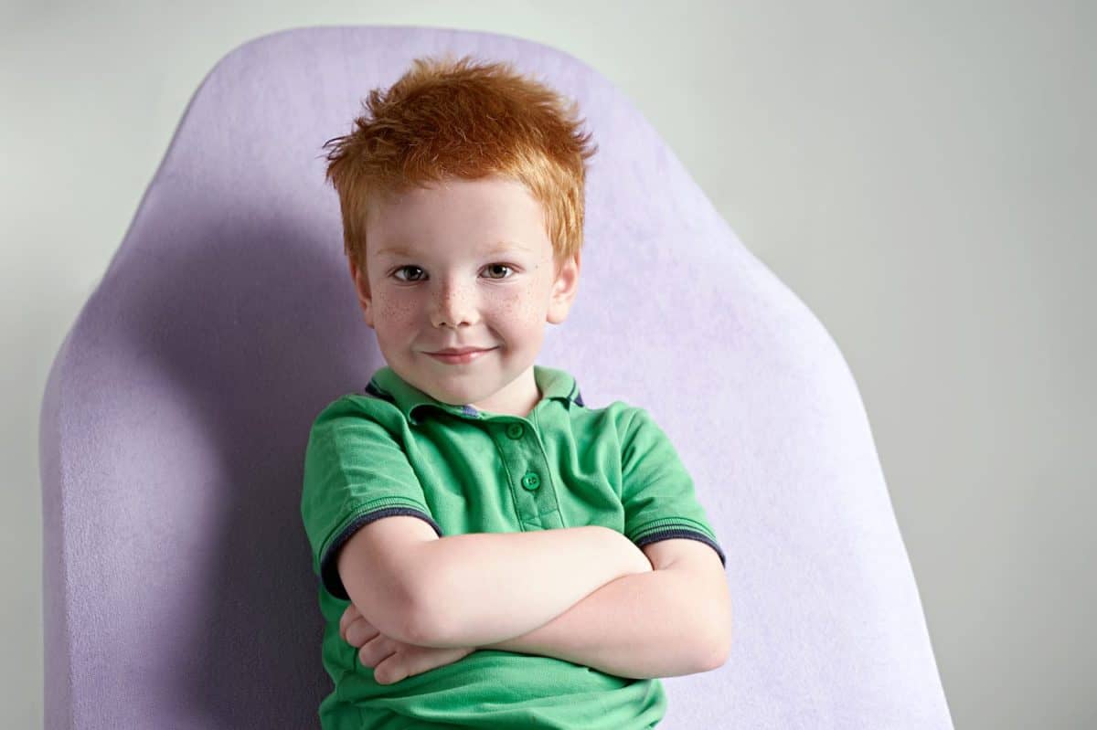 Cute red haired freckled little boy wearing green polo t-shirt sitting on a couch in doctor's office waiting for medical examination