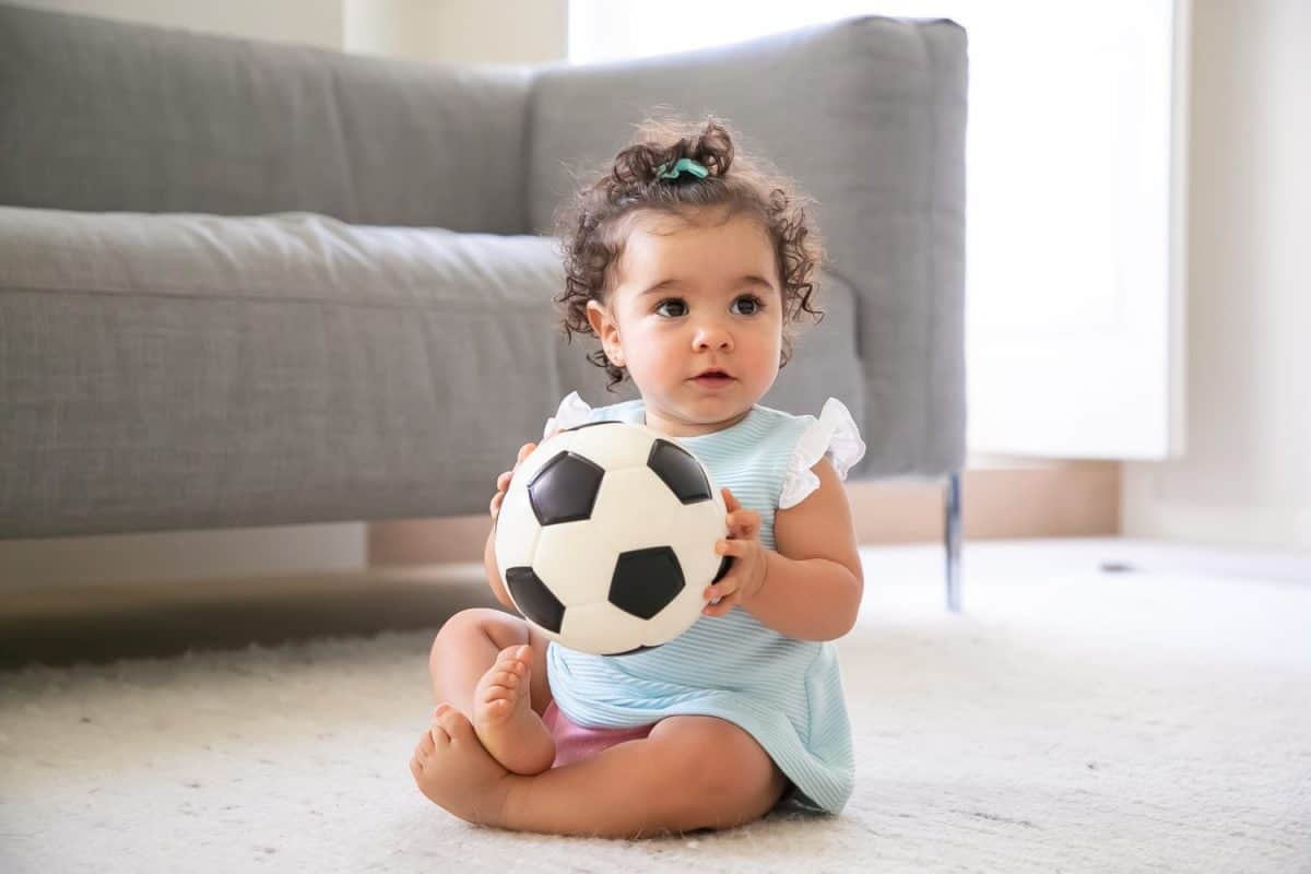 Adorable black haired baby girl in pale blue clothes sitting on floor at home, looking away, playing soccer ball. Kid at home and childhood concept