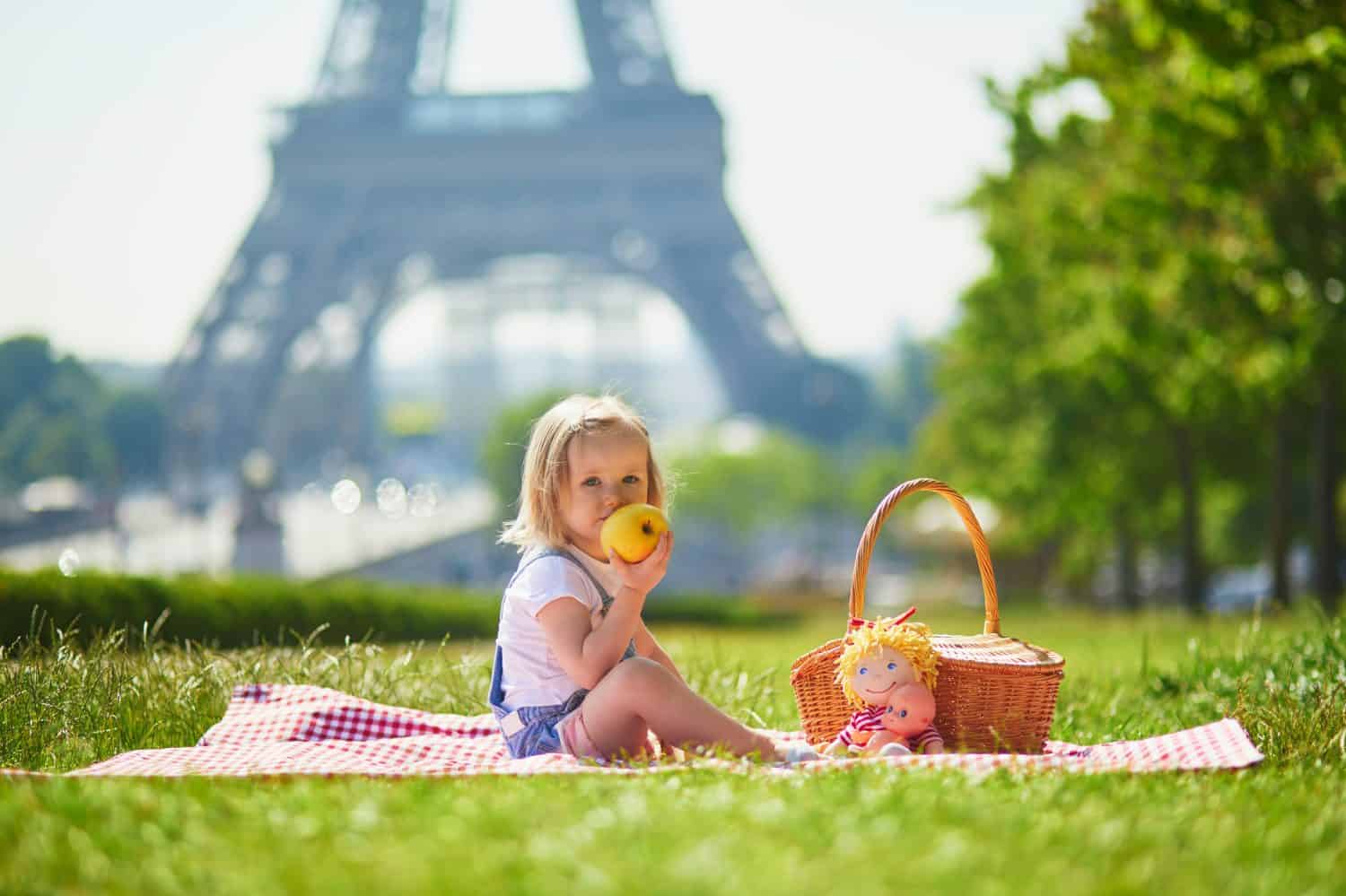 Cheerful toddler girl having picnic near the Eiffel tower in Paris, France. Happy child playing with toys in park on a summer day. Kid enjoying healthy snacks outdoors