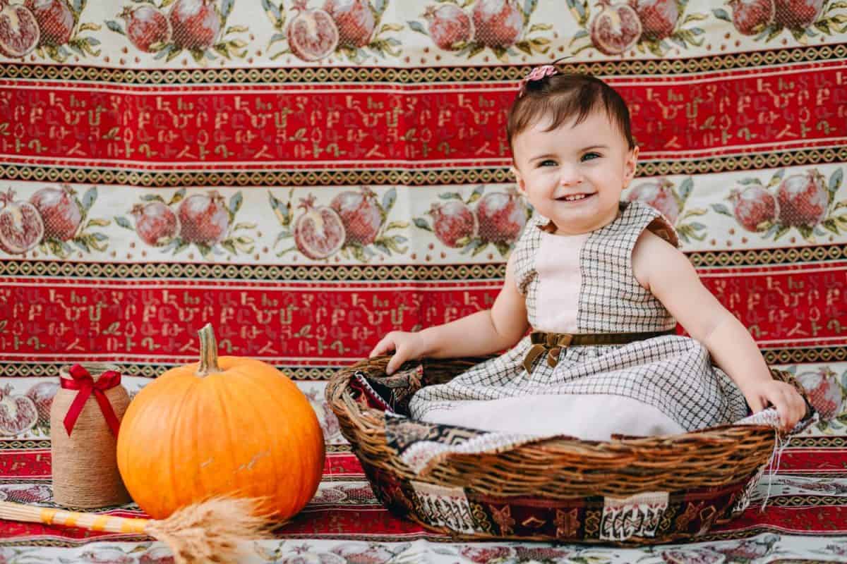 Funny armenian baby girl sitting in big straw basket and laughing next to the pumpkin