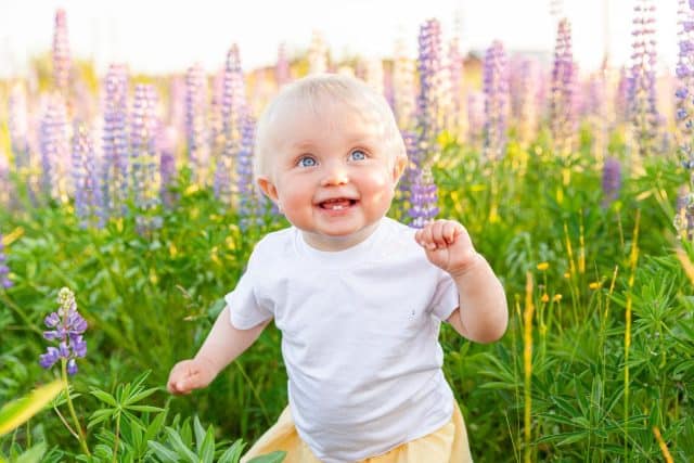 Happy little girl smiling outdoor. Beautiful blond young baby girl resting on summer field with blooming wild flowers green background. Free happy kid, childhood concept. Positive toddler child