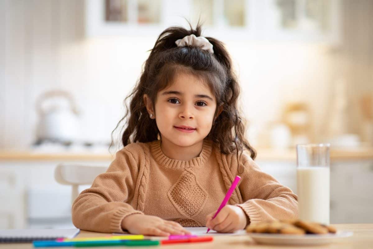 Beautiful Little Arab Girl Sitting At Table In Kitchen And Drawing, Cute Female Kid Painting With Pencils At Home, Enjoying Preschooler Development Activities And Looking At Camera, Copy Space