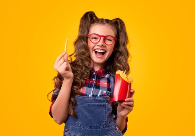 Optimistic stylish girl in glasses smiling for camera and eating french fries against yellow background