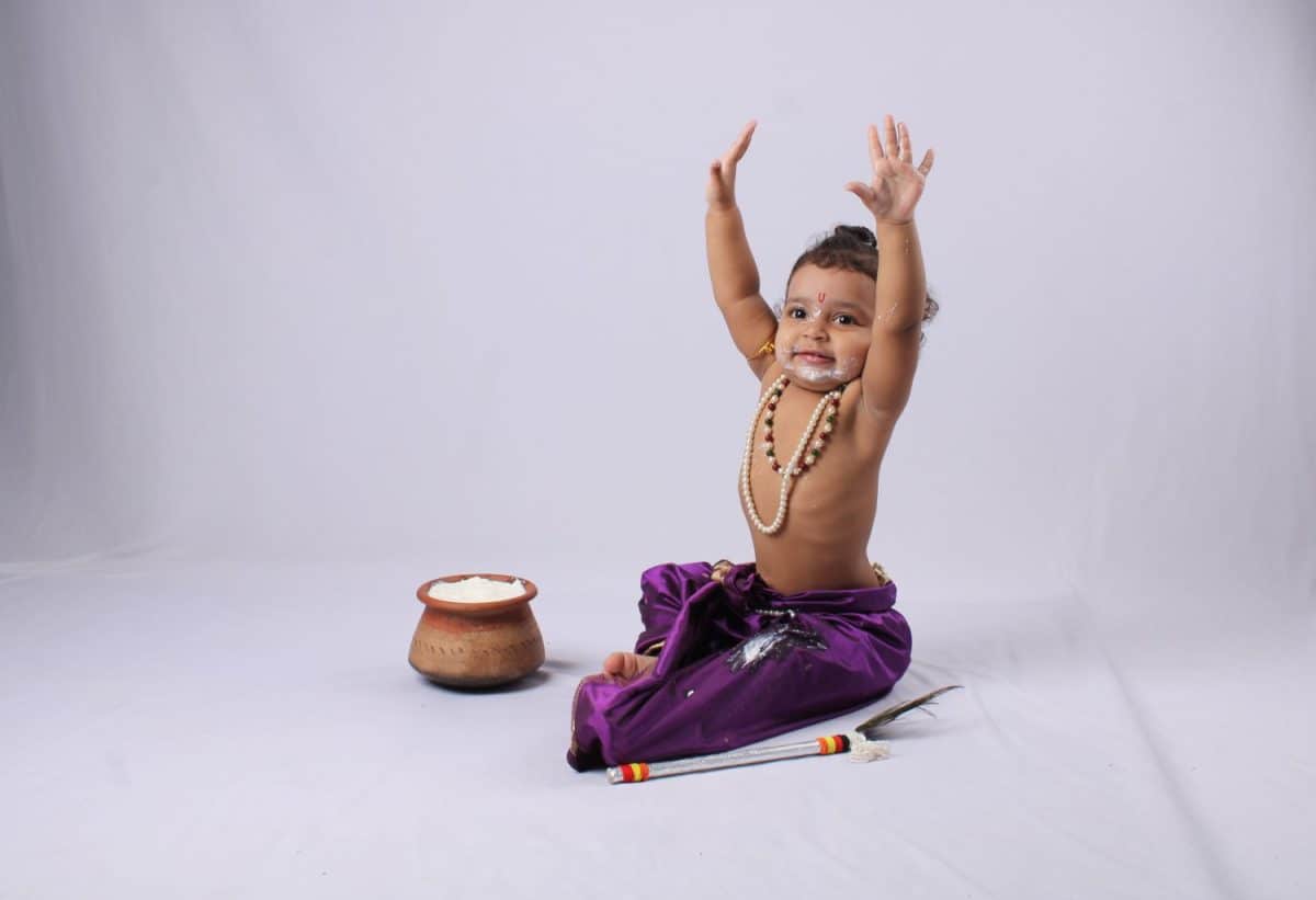 adorable Indian baby in krishna kanha or kanhaiya dress posing with his flute and dahi handi (pot with curd) on white background. sitting pose