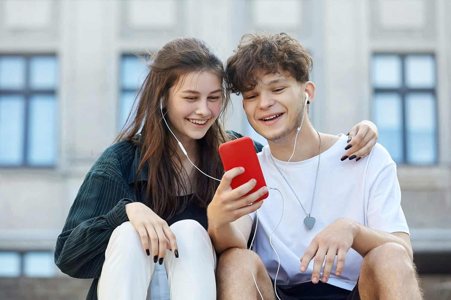 young guy and a girl sitting on the street, who have one pair of headphones for two, look into a red smartphone with a smile.