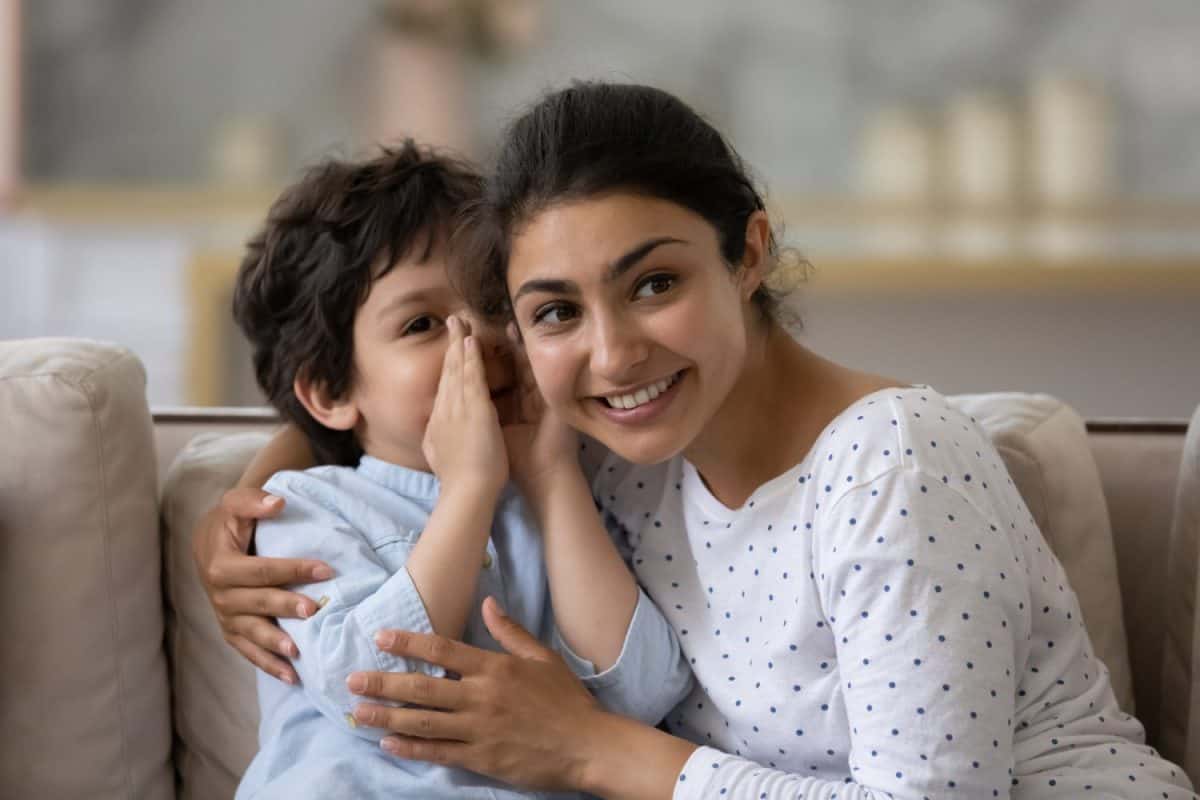 Cute little son telling secret to cheerful Indian mom, whispering in ear. Happy loving mother and kid enjoying being friendship, trust, spending leisure time together, having fun, talking at home