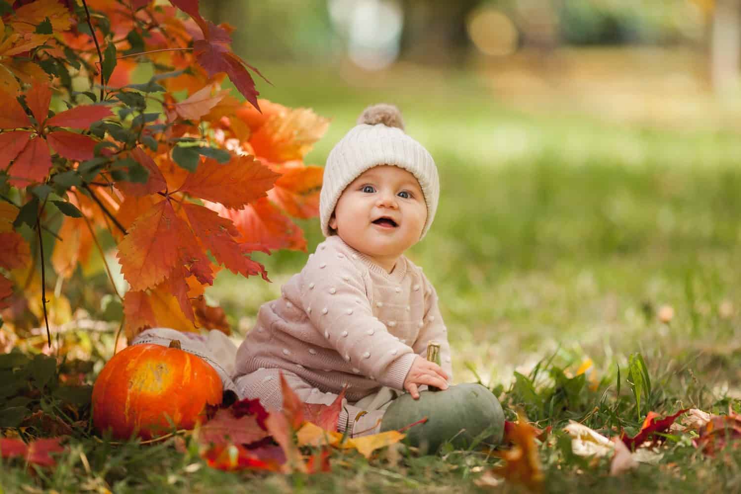 A cute little adorable baby girl in puff ball hat is sitting on the ground in the park with a pumpkin smiling and Playing. Happy childhood concept. looking at the camera