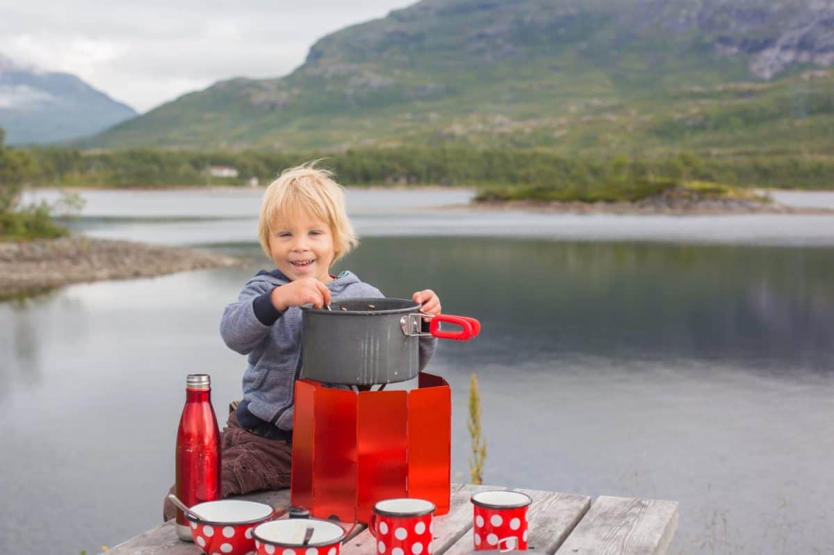 Happy child, eating on a bench on a rest stop along the road, mom cooking on small camping cooker and having family lunch on the road