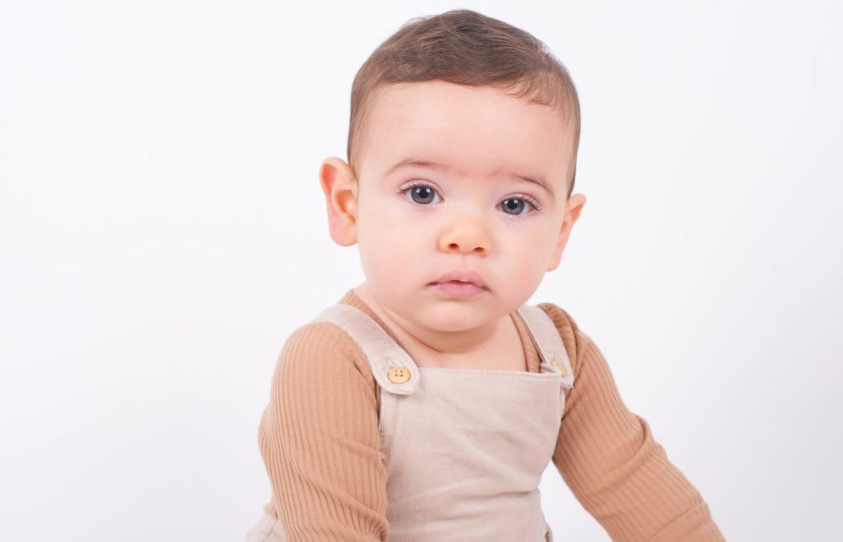 Close up portrait of adorable seven month old baby boy wearing beige overalls sitting on white background and looking to the camera front view. Childhood and innocence concept.