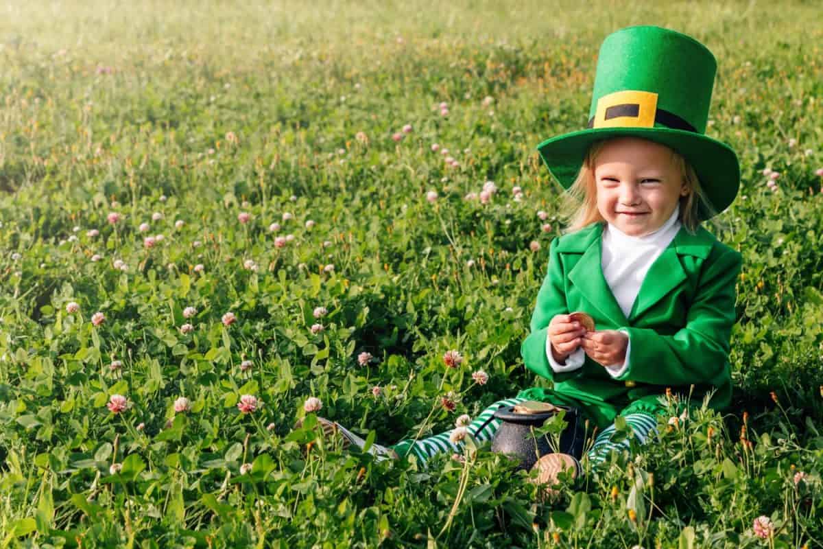 St. Patrick's Day. A girl in a green suit is sitting in a large clearing of clover, holding a pot with gold coins in her hands. copyspase. Happy joyful toddler baby