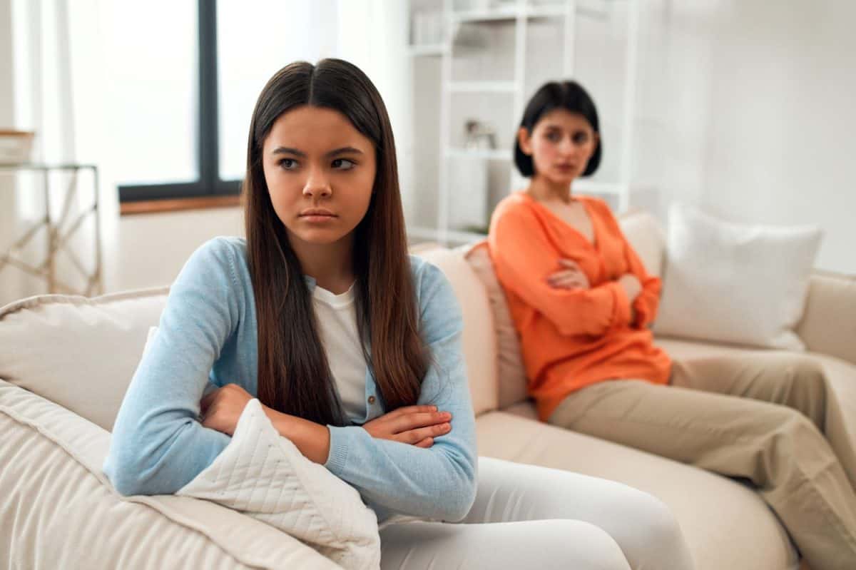 Upset mother and daughter sitting back to back on the couch, avoiding talking and staring, mom and teenage girl fighting or arguing, stubborn parent and child refusing to compromise.