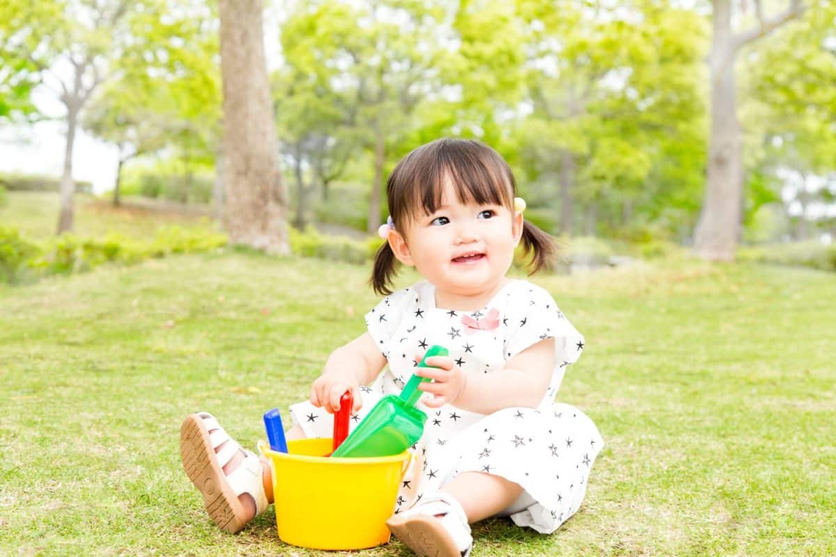 Little girl sitting on grass against backdrop of fresh green trees and playing alone with toys. Image of early childhood, playing alone, childcare