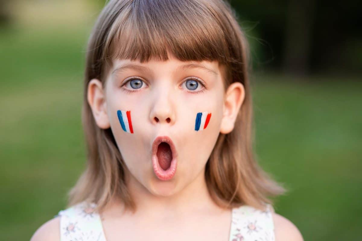 Portrait of surprised 7 years old child with painted cheeks in flag of France walking outdoor. Cute little girl screaming with opened mouth and crazy expression looking at camera.