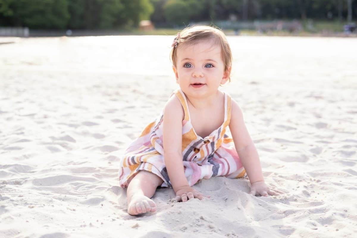 happy baby girl sitting on the beach on sand wearing striped summer dress on sunny day