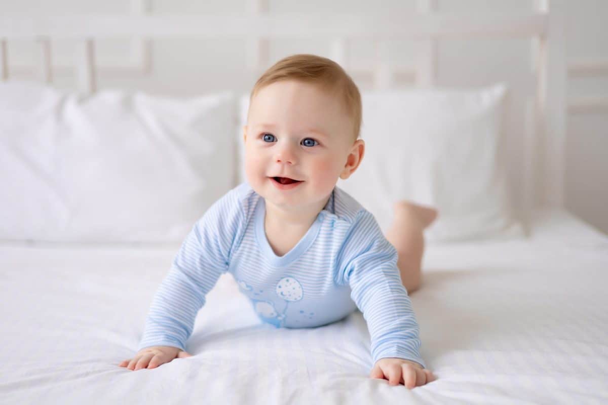 a cute healthy little baby is lying on a bed on white bedding at home in a blue bodysuit. The kid looks at the camera, smiles