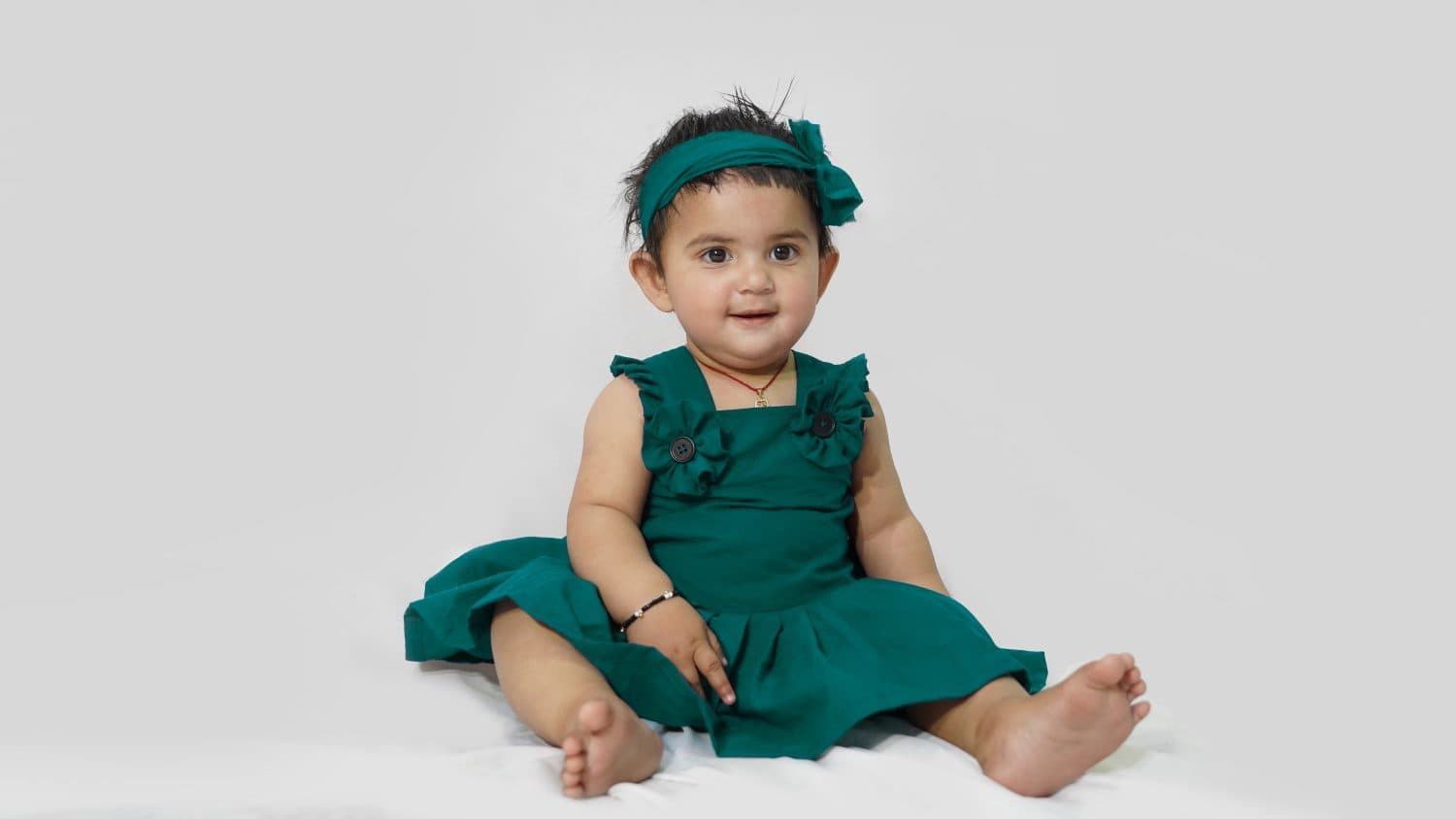 Indian baby girl sitting on white background, looks beautiful in dark green frock