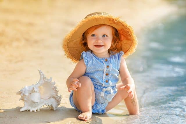 Red hair baby girl in blue clothes and a straw hat sits on the sand on the beach in summer. The shell is nearby the kid.