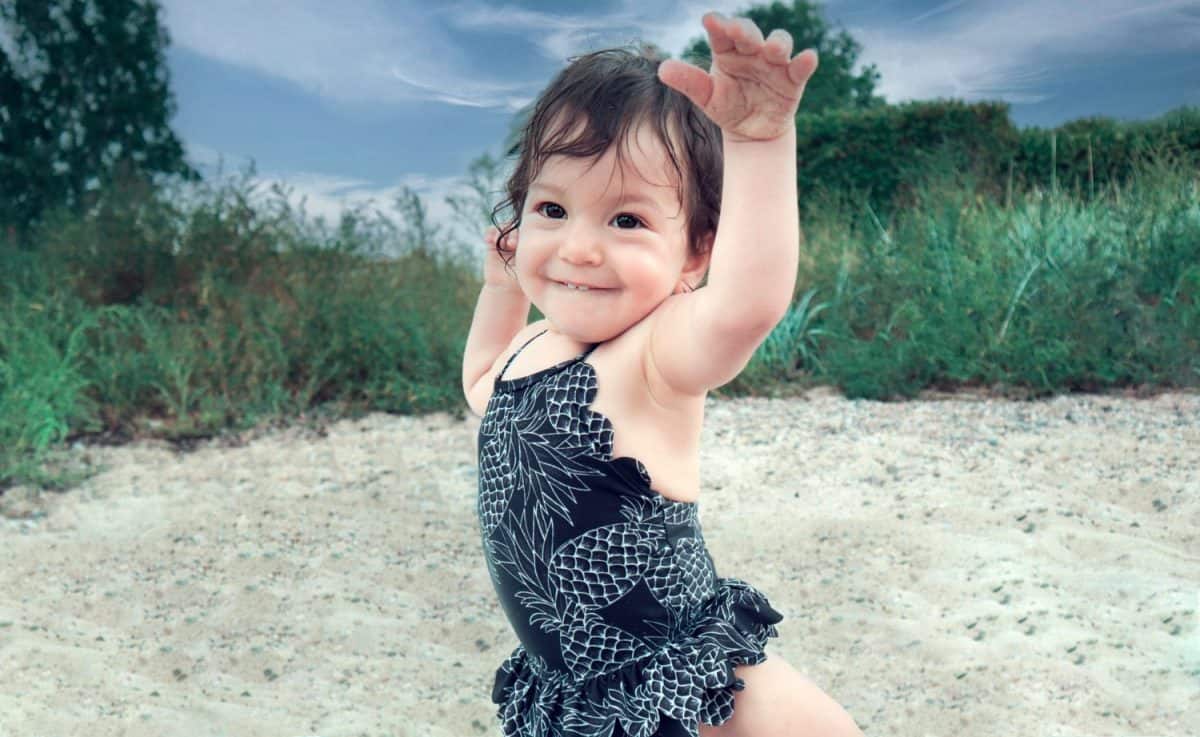 Happy, smiling, adorable brunette baby girl with dark brown eyes on the sandy beach wearing black swimsuit and holding her hands up on a family holiday.