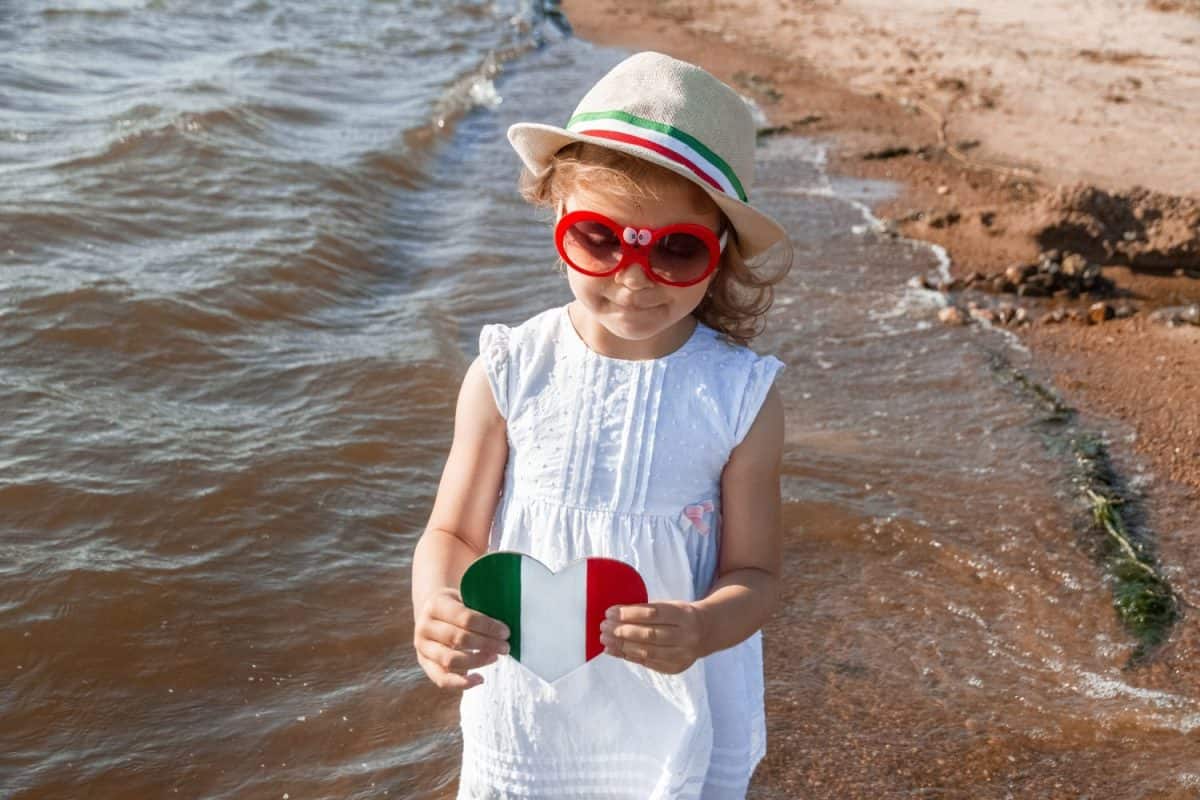 Girl of 3 years in white dress in red glasses, hat and wooden heart with colors of italy flag on beach summer, selective focus