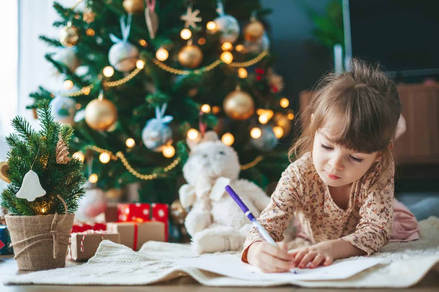 Cute little child girl writing letter to Santa Claus or writing dreams of a gift with near Christmas tree. Merry Christmas and Happy New Year!