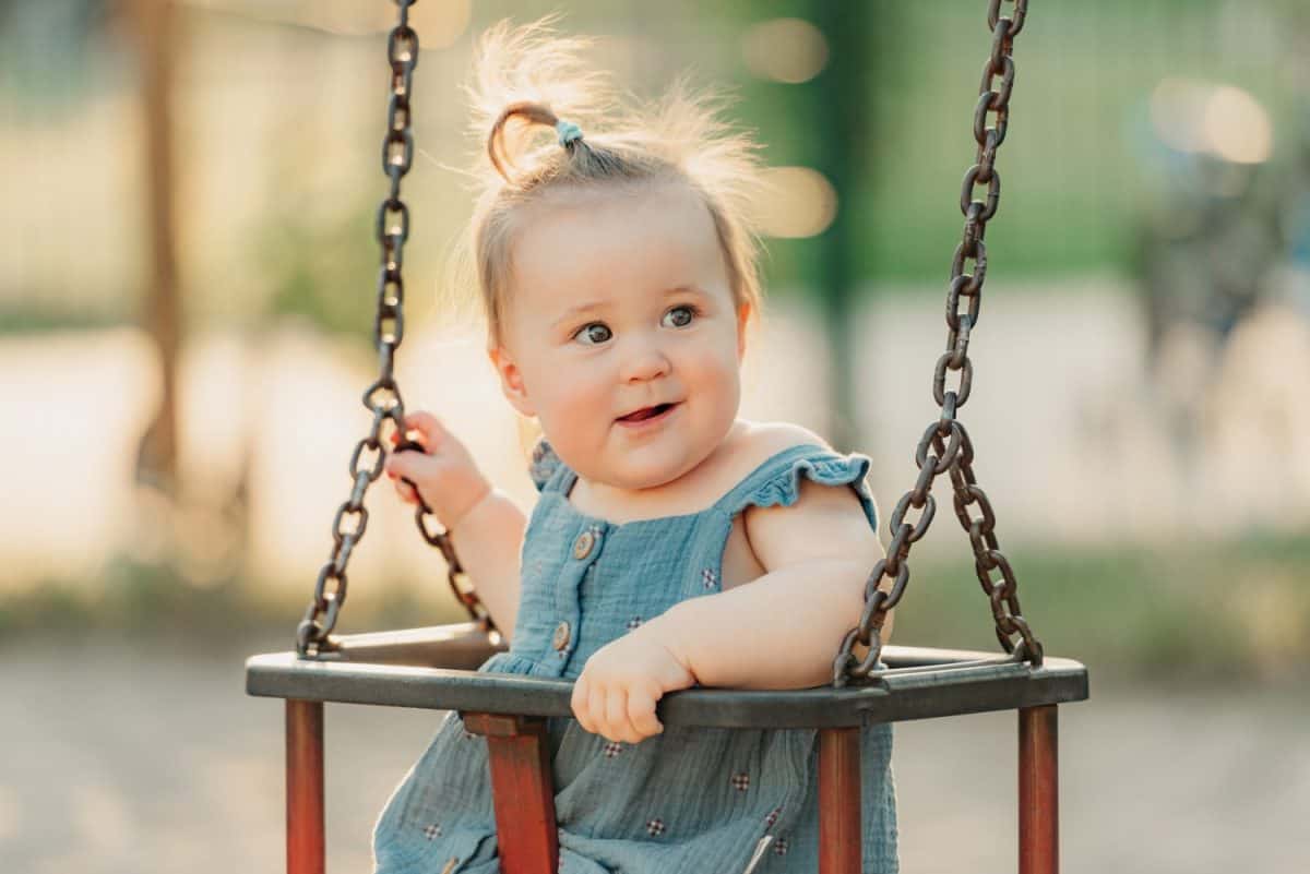 A happy toddler baby girl in a dress on a swing on the warm summer evening