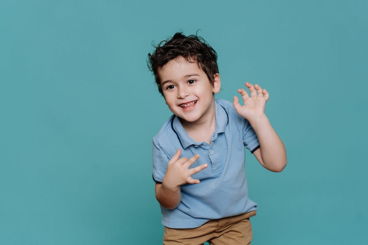 Happy curly little Italian boy in blue polo dancing, smiles against turquoise studio background. Healthy caucasian toddler enjoying life. Childhood, entertainment for kids. Caucasian baby boy smiles.