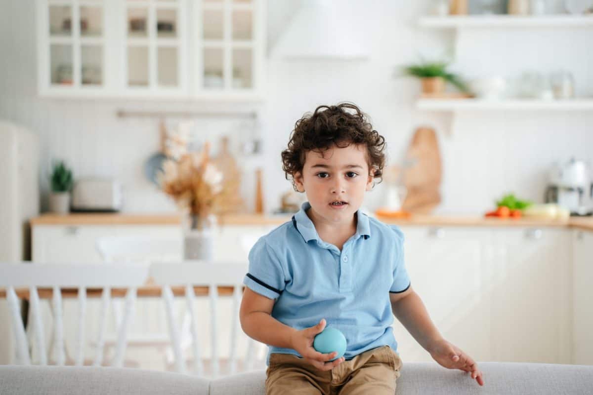 Cute handsome curly Italian boy sitting on couch at home against blurry kitchen holds little ball. Playful caucasian toddler playing alone indoors. Childhood, smart kids. Hispanic baby looks at camera