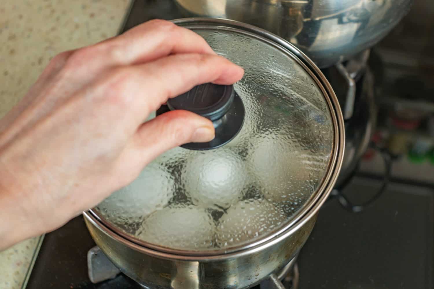 Pot of boiling water in which white chicken eggs are boiled. Water boils and boils in pan with boiled eggs. Female hand closes pan with boiled white chicken eggs with transparent glass roof. Close-up.