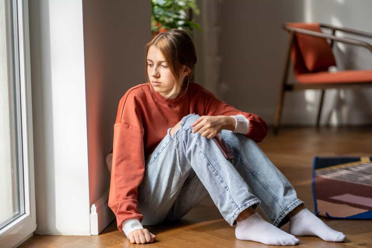 Upset teen girl sit on floor sadly look out window worried about teenage problem at school and communication with parent. Worried girl tensely suffer about bullying at school, unrequited love with boy