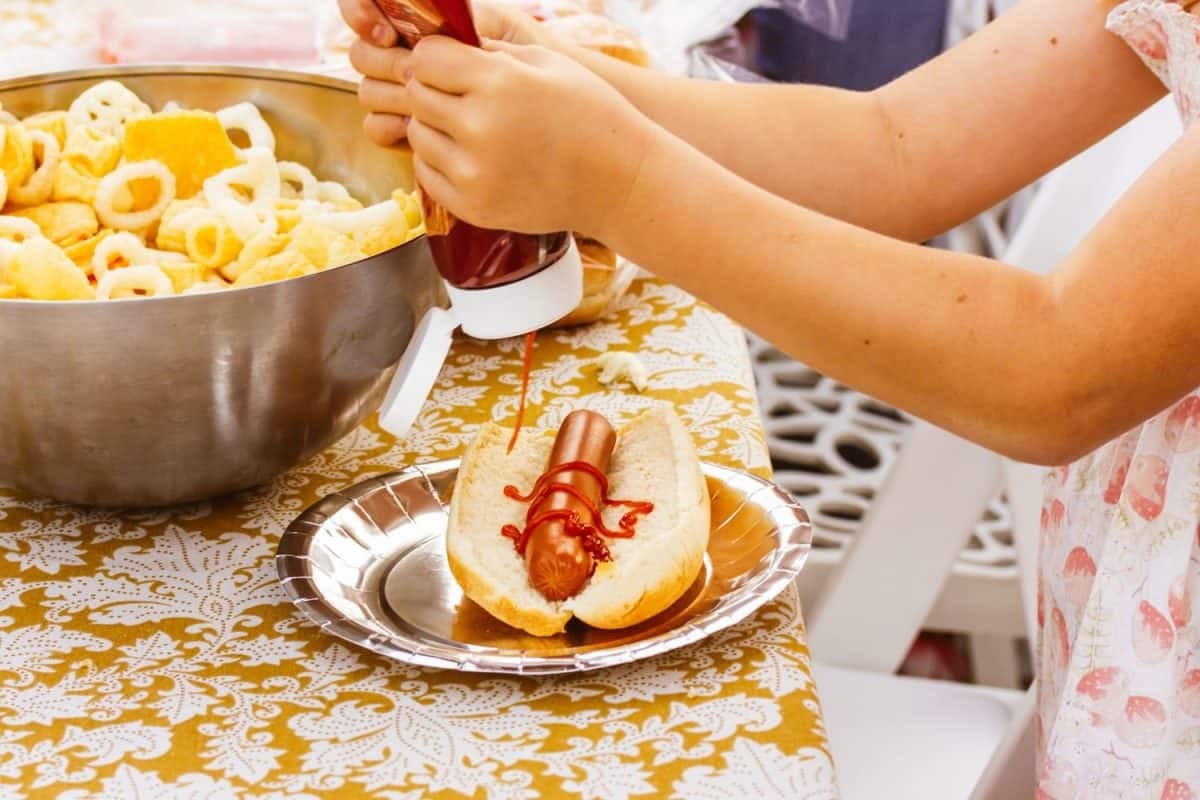 A little girl squeezing ketchup on a hotdog. Picnic, food, buffet table with beautiful tablecloth. Child eating American fast food, junk food, tomato catsup, snacks outside. A child, kid eats outdoor.