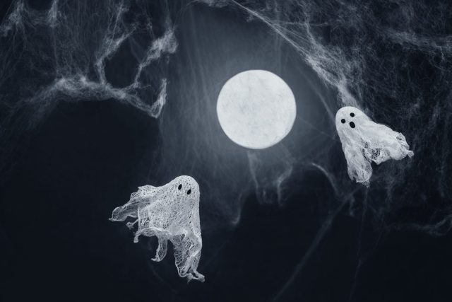 Ghosts of gauze on a dark background with glowing moon and spider web. Creative DIY Halloween decor. Halloween greeting card. Copy space.